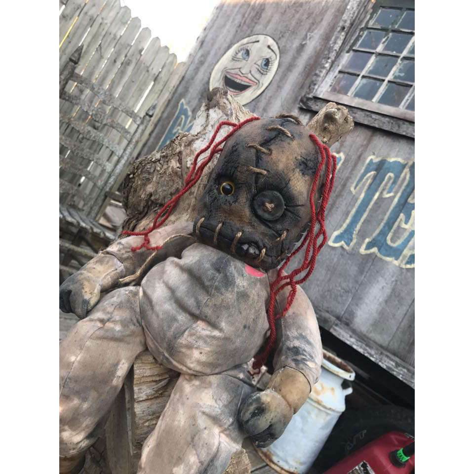 A worn and creepy cloth doll with red yarn hair, stitched facial features, and an eye patch, sitting against a rustic background with a wooden fence and a painted smiley face.